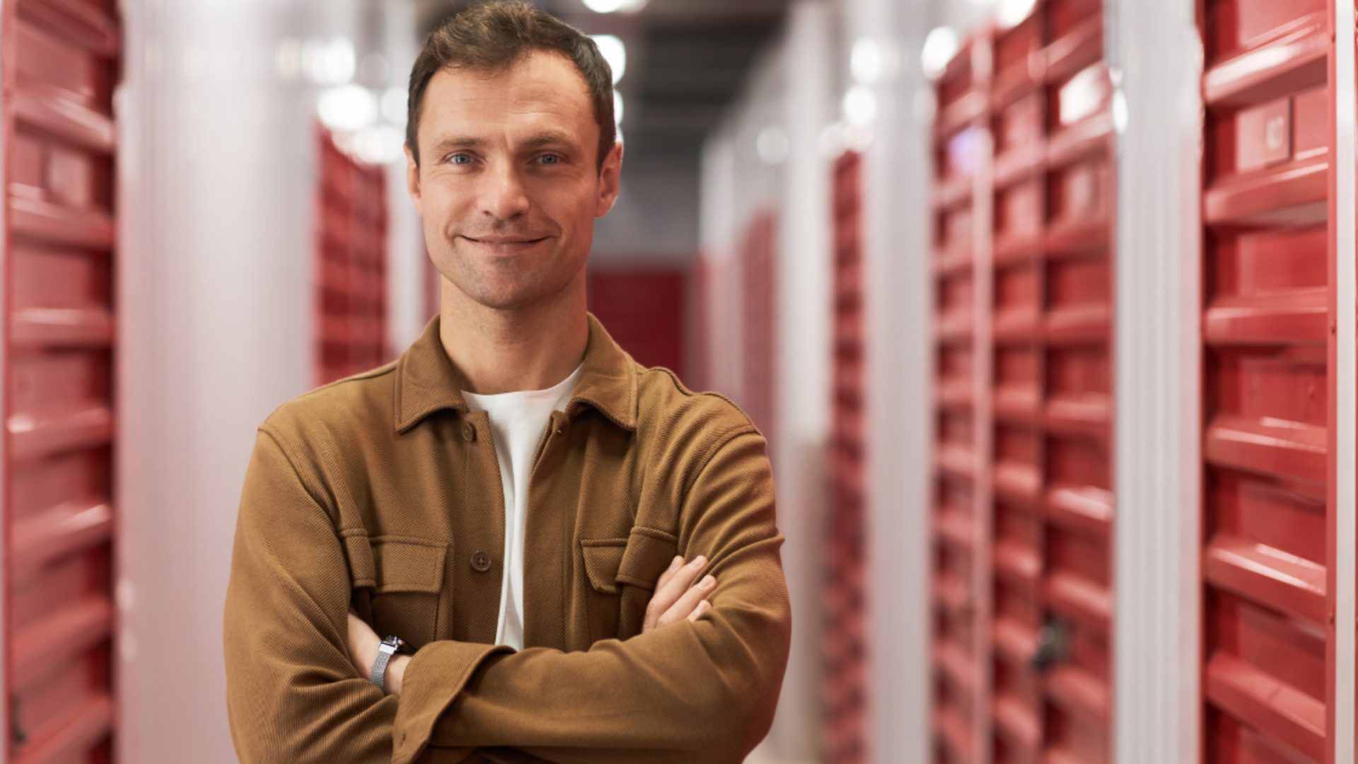 A smiling person with arms crossed, standing in a hallway lined with red self-storage unit doors.