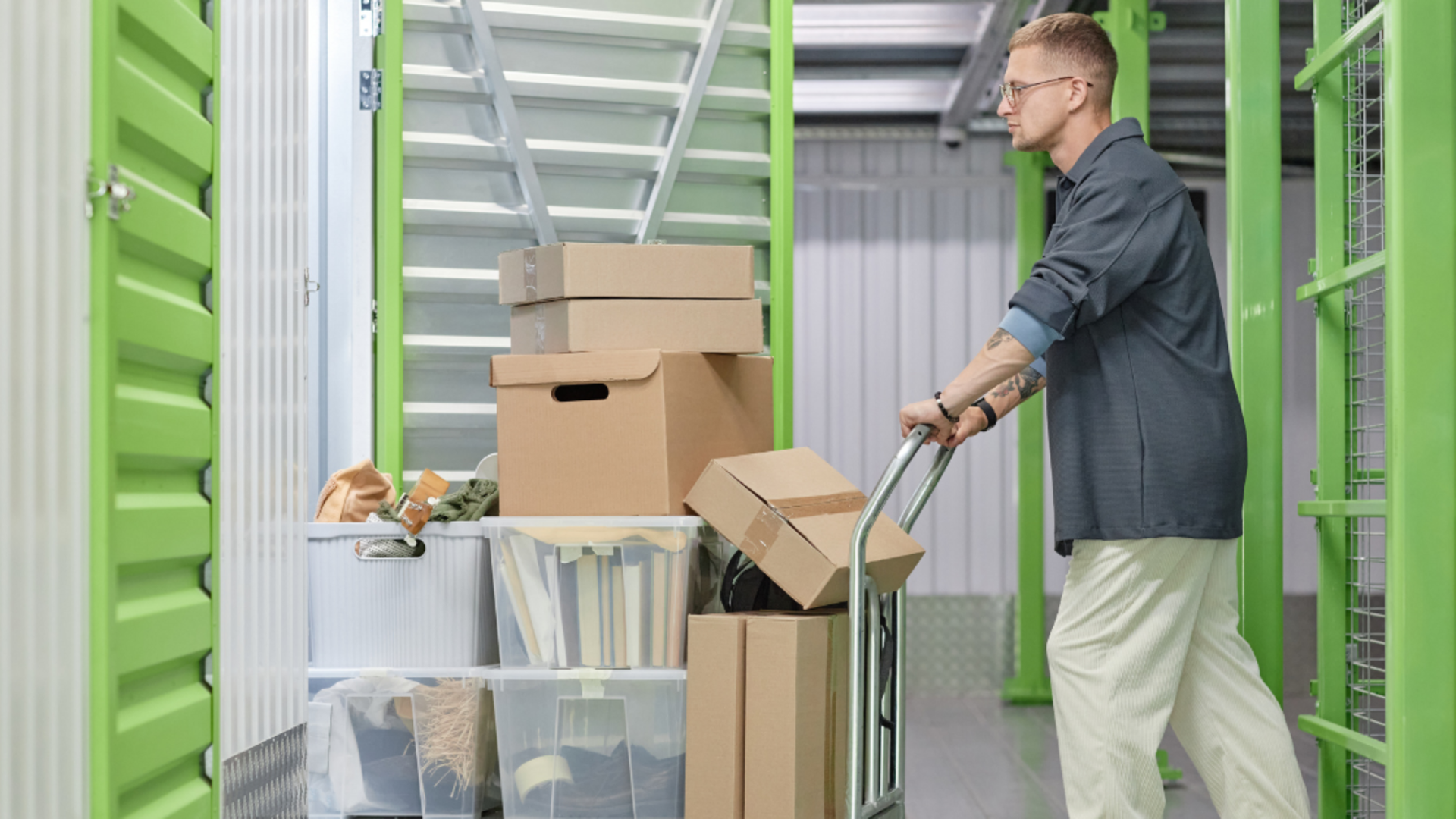 A person in a gray shirt and light pants pushes a metal cart stacked with cardboard boxes and storage bins in a facility.
