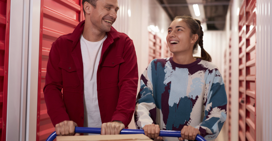 A man and woman smile while pushing a cart filled with cardboard boxes down a bright hallway lined with red storage units.