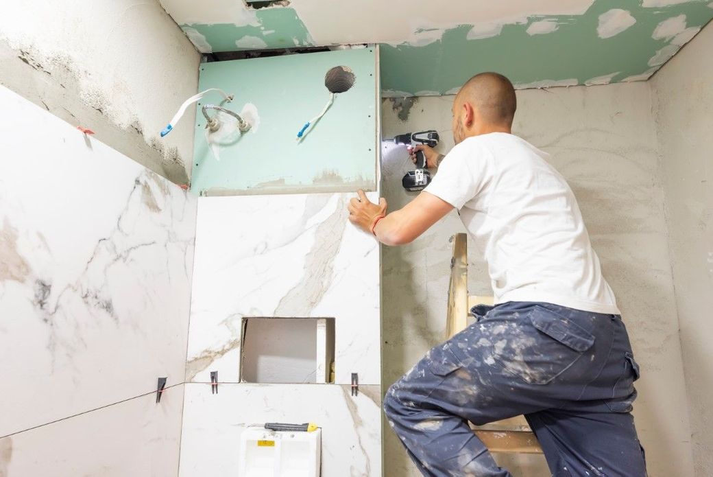 Contractor installing large wall tiles during a bathroom remodel, fitting marble-look panels around a shower niche and plumbing fixtures.