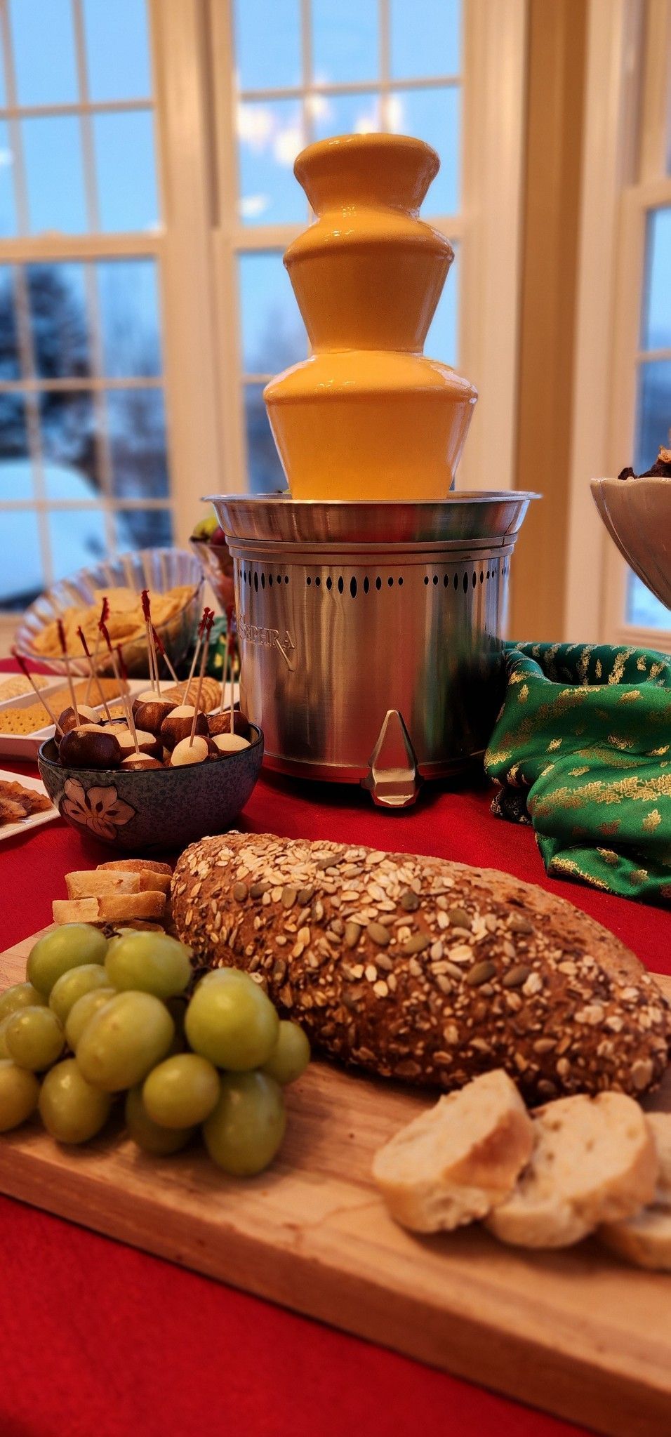 A chocolate fountain is sitting on a table next to a cutting board with grapes and bread.