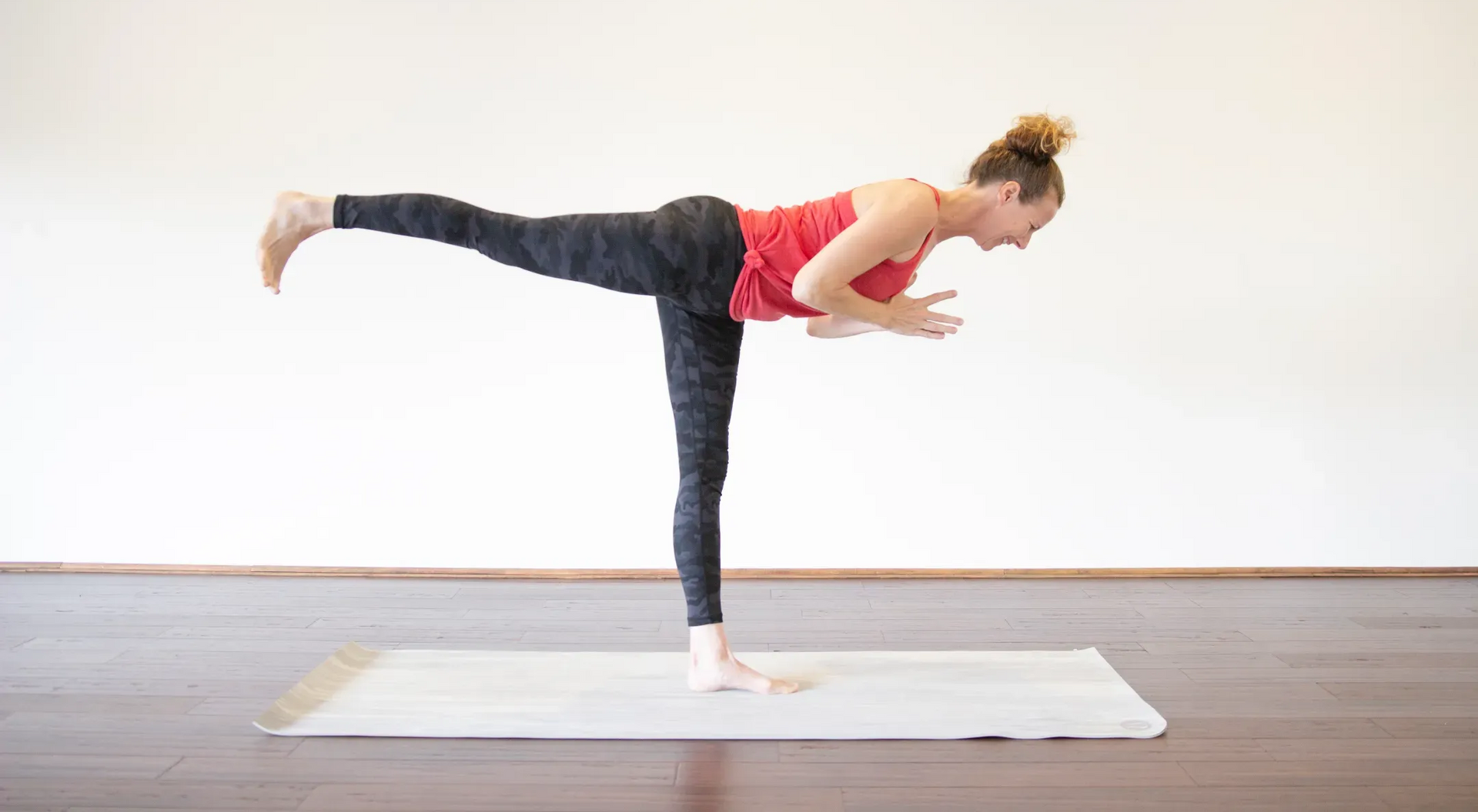 Woman in Warrior III yoga pose, balanced on one leg, arms clasped, in a studio.