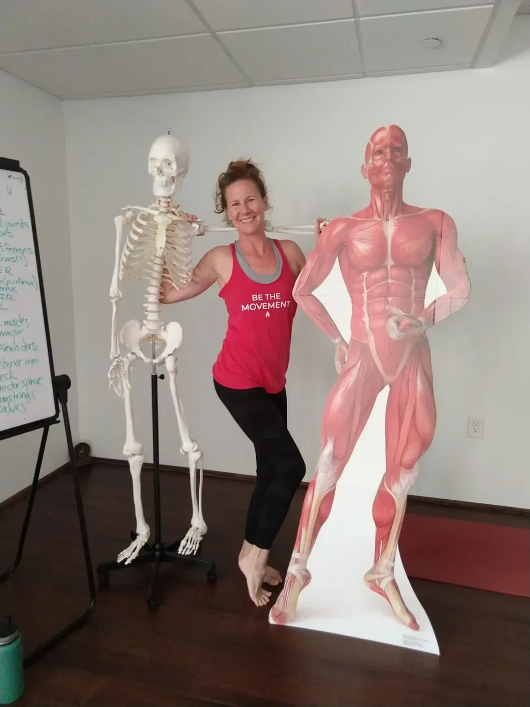 Woman poses with a skeleton and muscle man cutouts in a yoga studio.