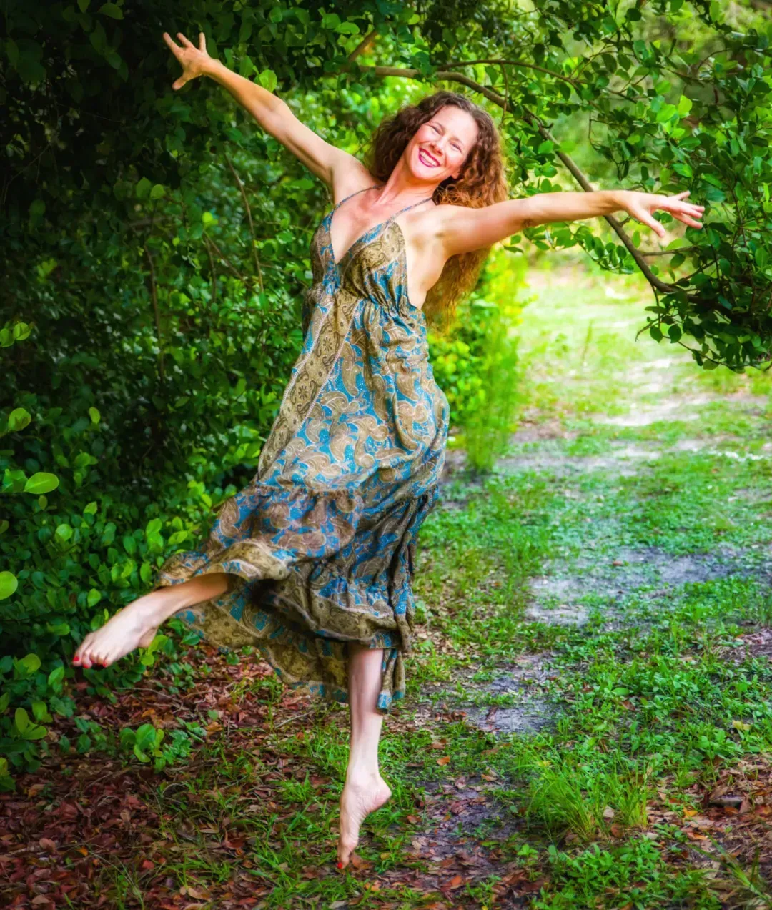 Woman in flowing dress leaps joyfully on a path lined with lush greenery.