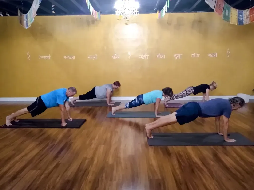 People in a yoga studio, practicing planks on mats. Gold wall, wood floor.