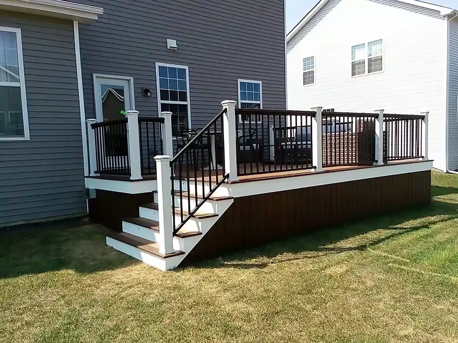 Brown deck with white trim and black railing, attached to gray house with grass lawn.
