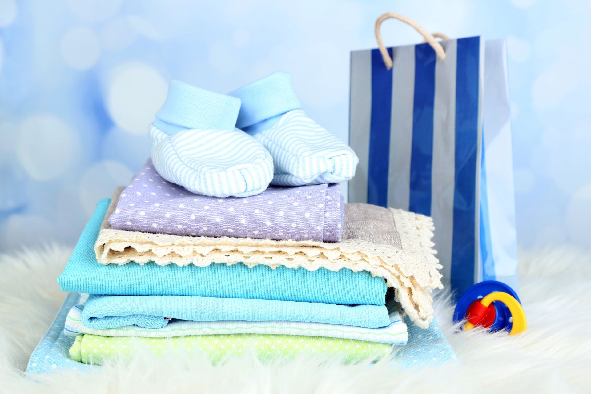 Pila de ropa de bebé con patucos, bolsa de regalo y juguete sobre una superficie esponjosa.