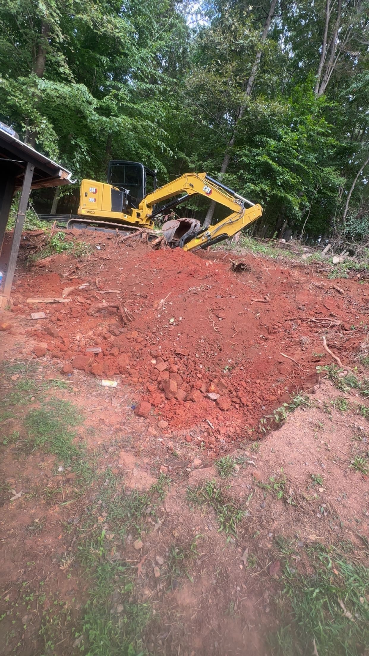 Yellow excavator on a red dirt pile, with trees in the background.