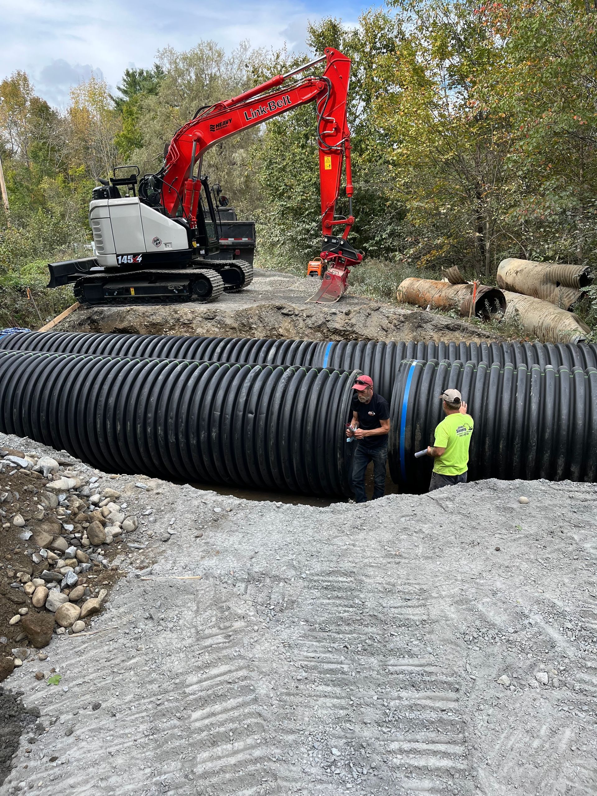 Two workers stand beside large corrugated black plastic pipes at a construction site with an excavator in the background.