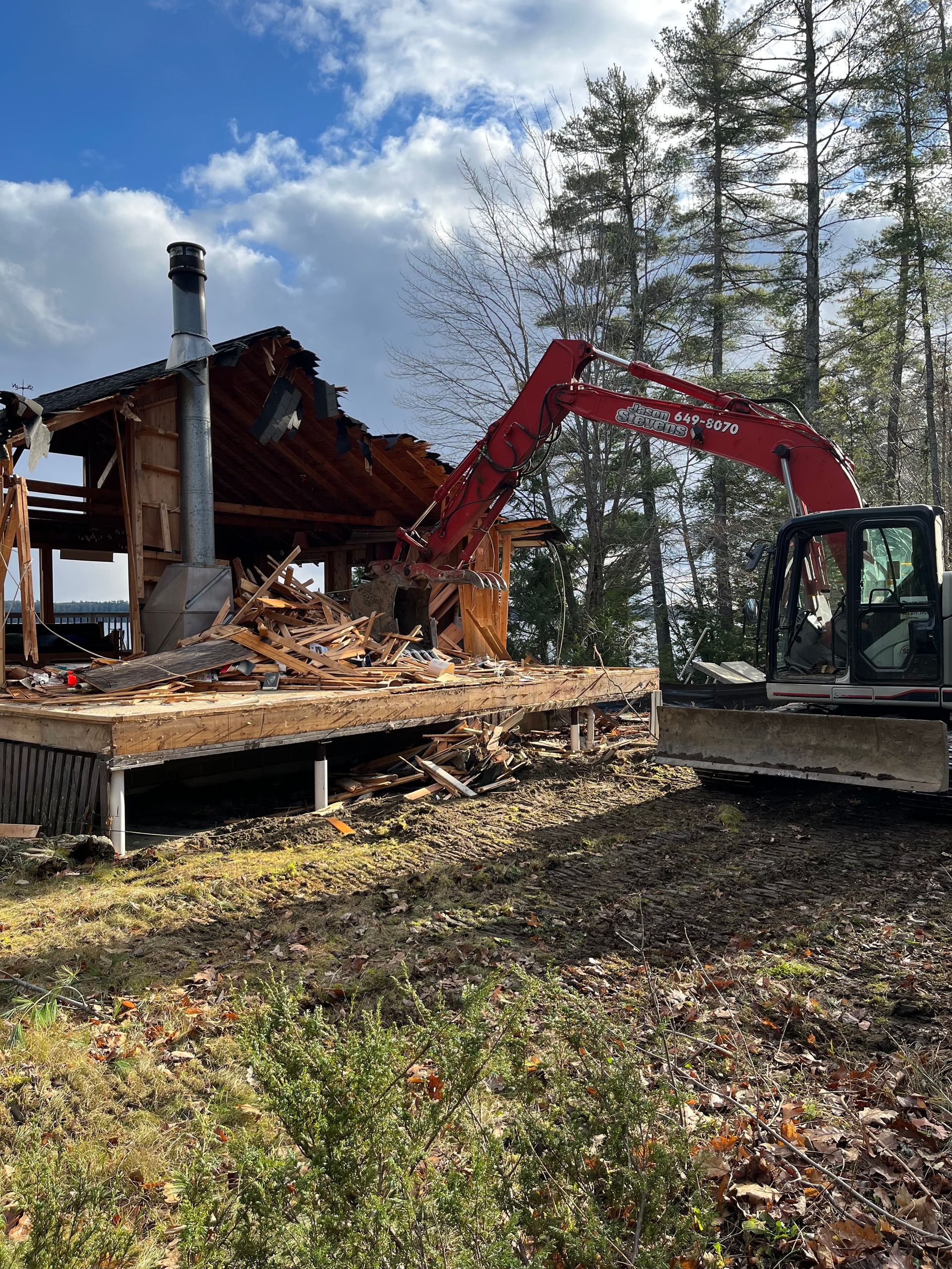 A red excavator demolishes a wood-framed structure near trees under a cloudy sky.