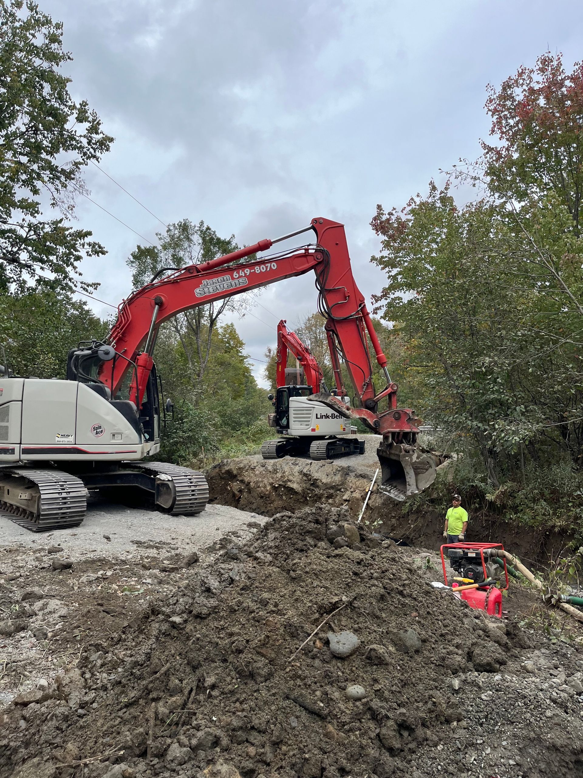 Two red excavators work on a construction site with piles of dirt and a worker in a high-visibility vest nearby.