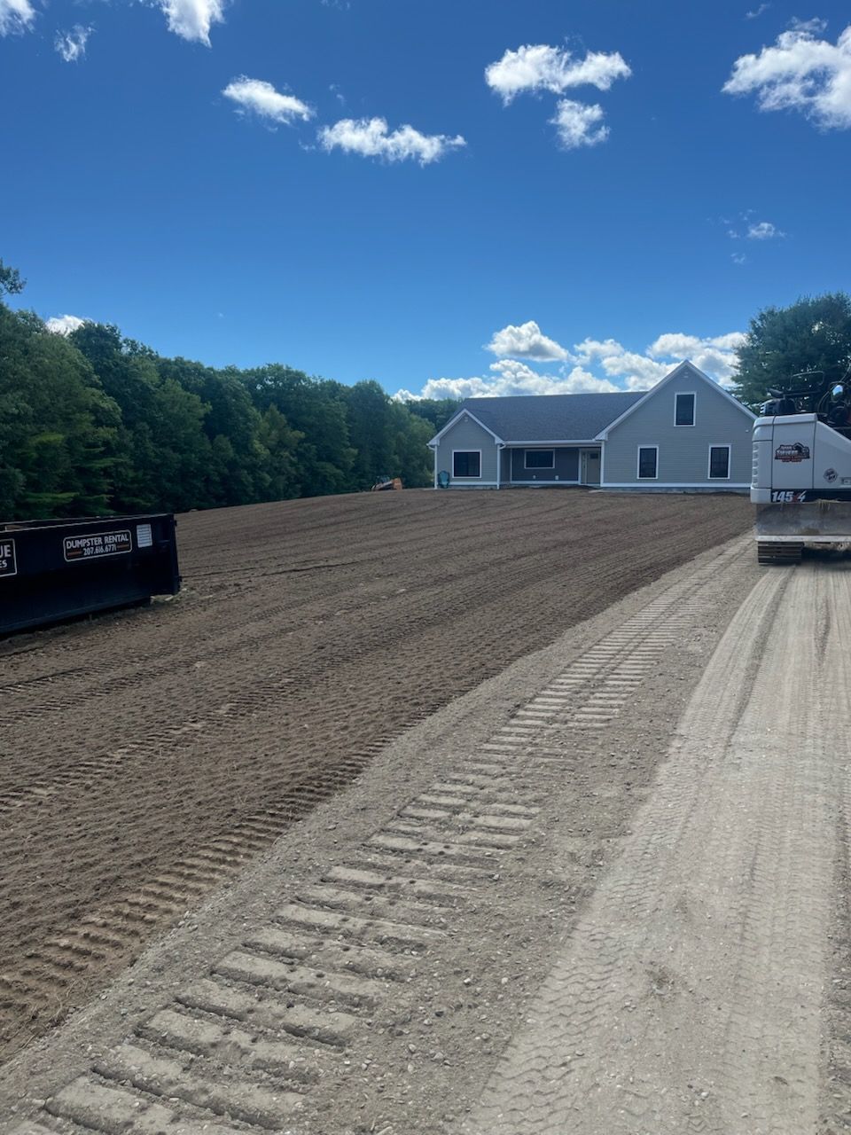 A graded dirt lot leading to a light-gray house under a blue, cloudy sky, with construction tracks visible in the soil.