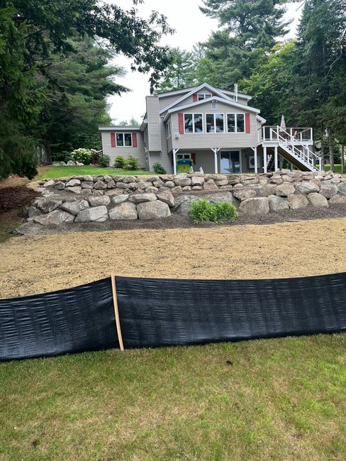 A multi-level lakeside home with stacked stone retaining walls and a black silt fence in the foreground.