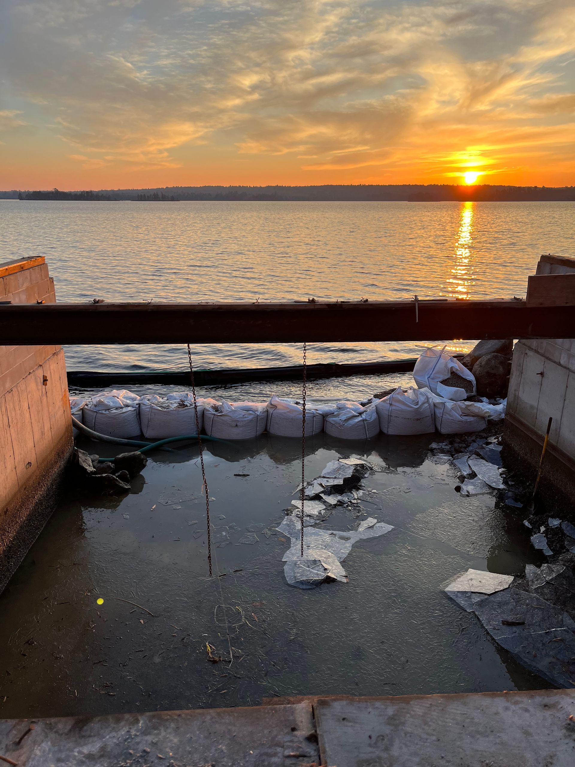 A sunset over water, viewed past a metal railing and a row of sandbags protecting a small, muddy area from the tide.