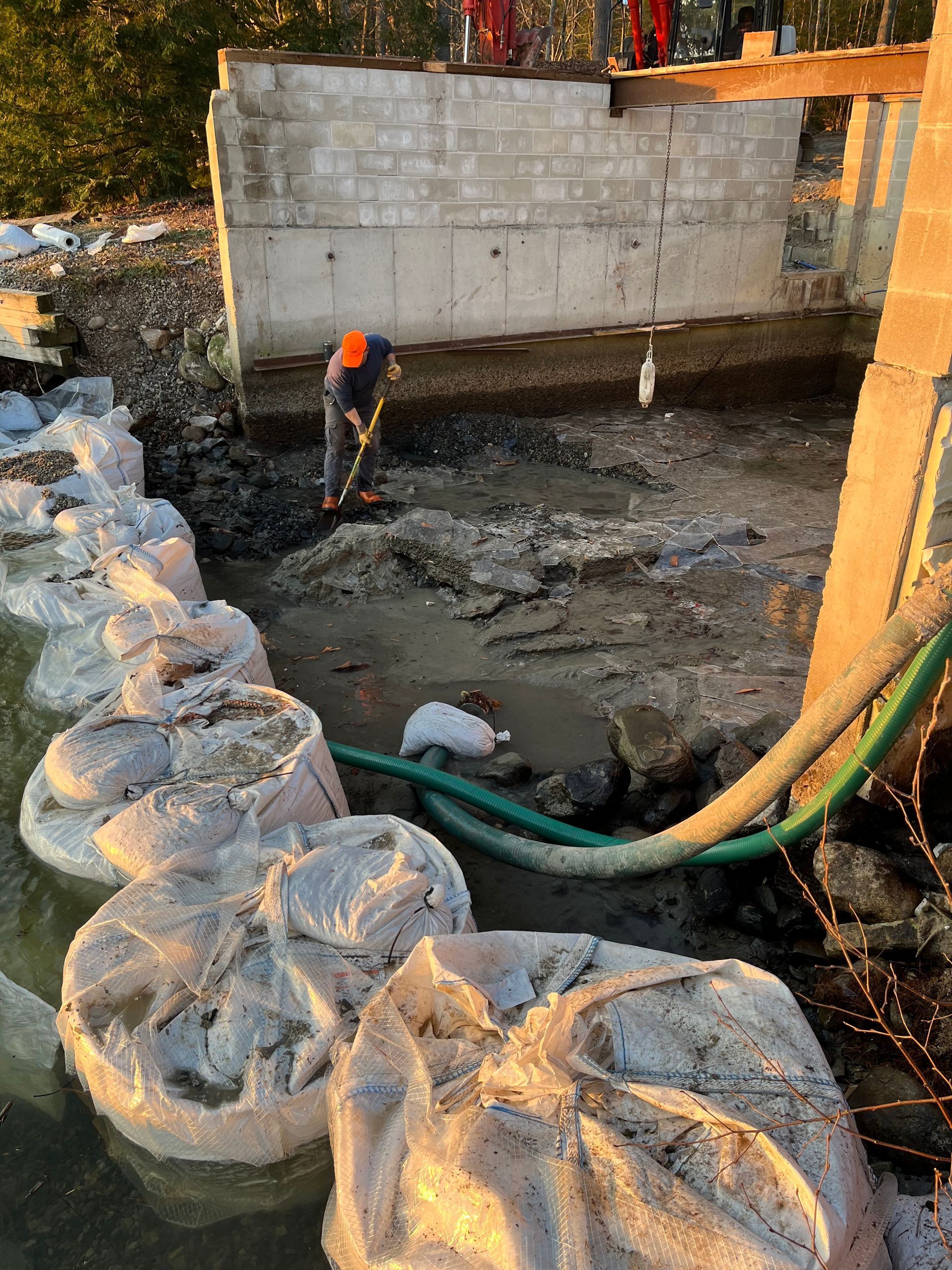 A worker in a high-visibility vest clears mud near a concrete wall foundation, flanked by large white sandbags and hoses.