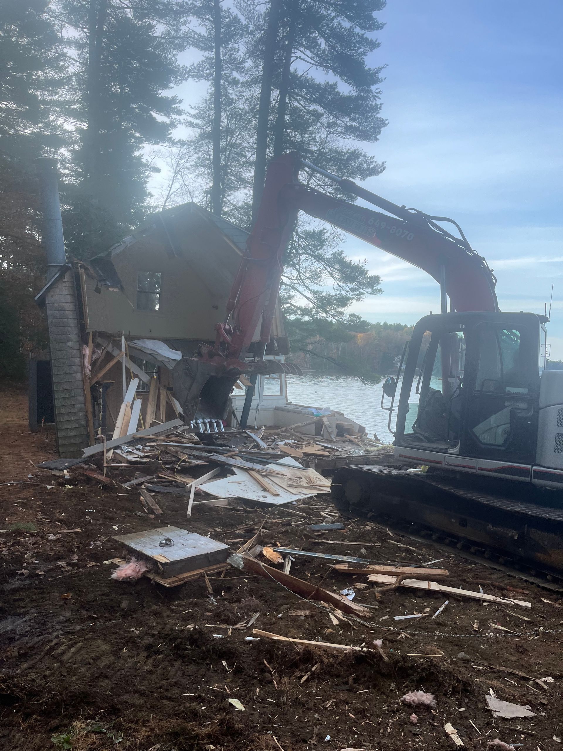 A large excavator tears down a wooden building near a lake shore surrounded by trees.