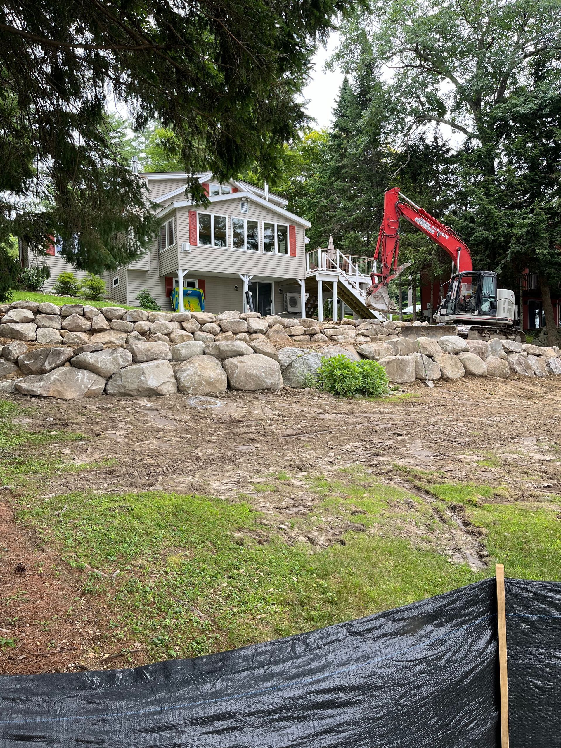 A house with a large stone retaining wall and a red excavator working in the backyard.
