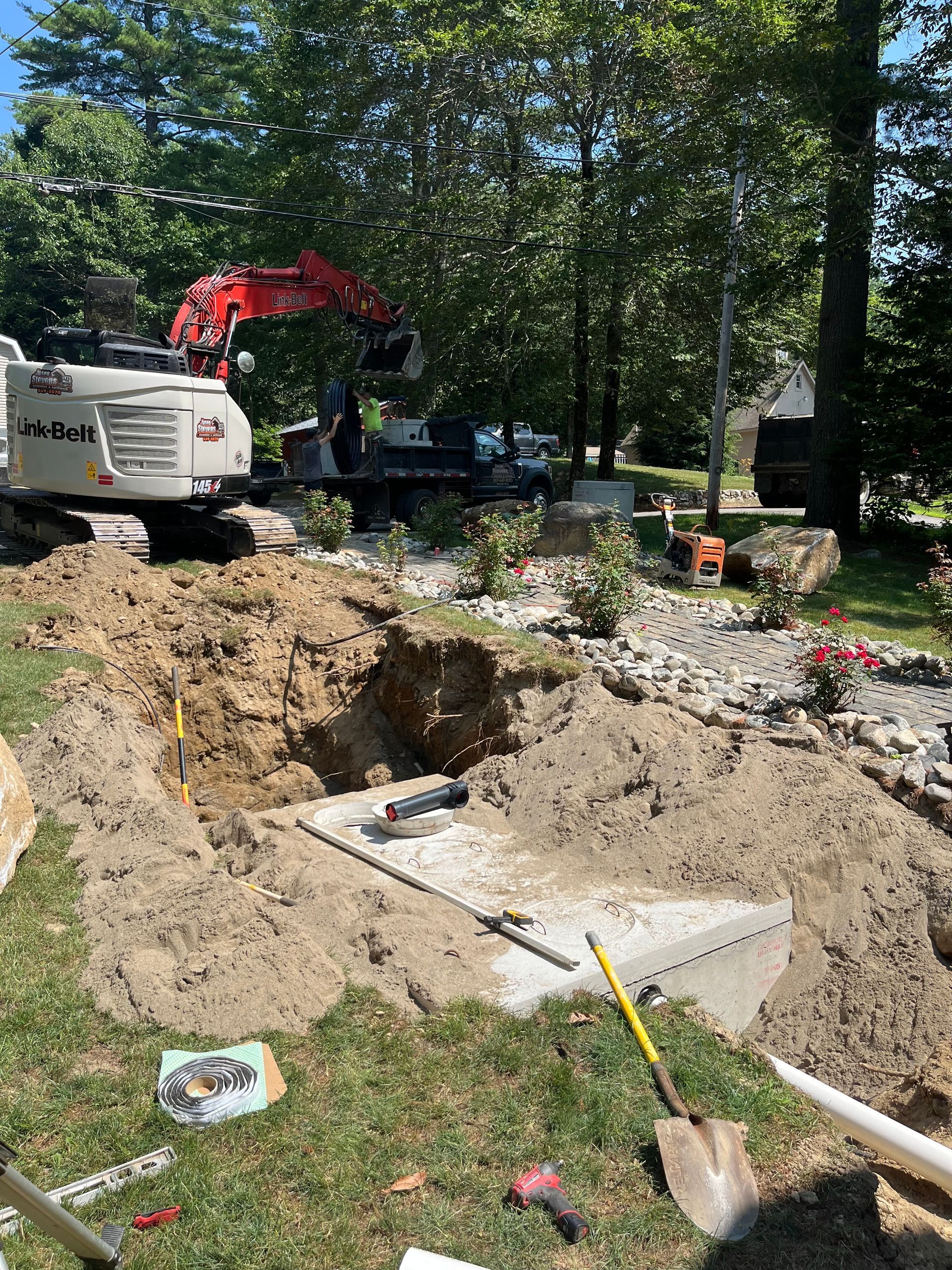 A red excavator sits in a grassy yard behind a large excavated trench containing a concrete septic or utility tank.