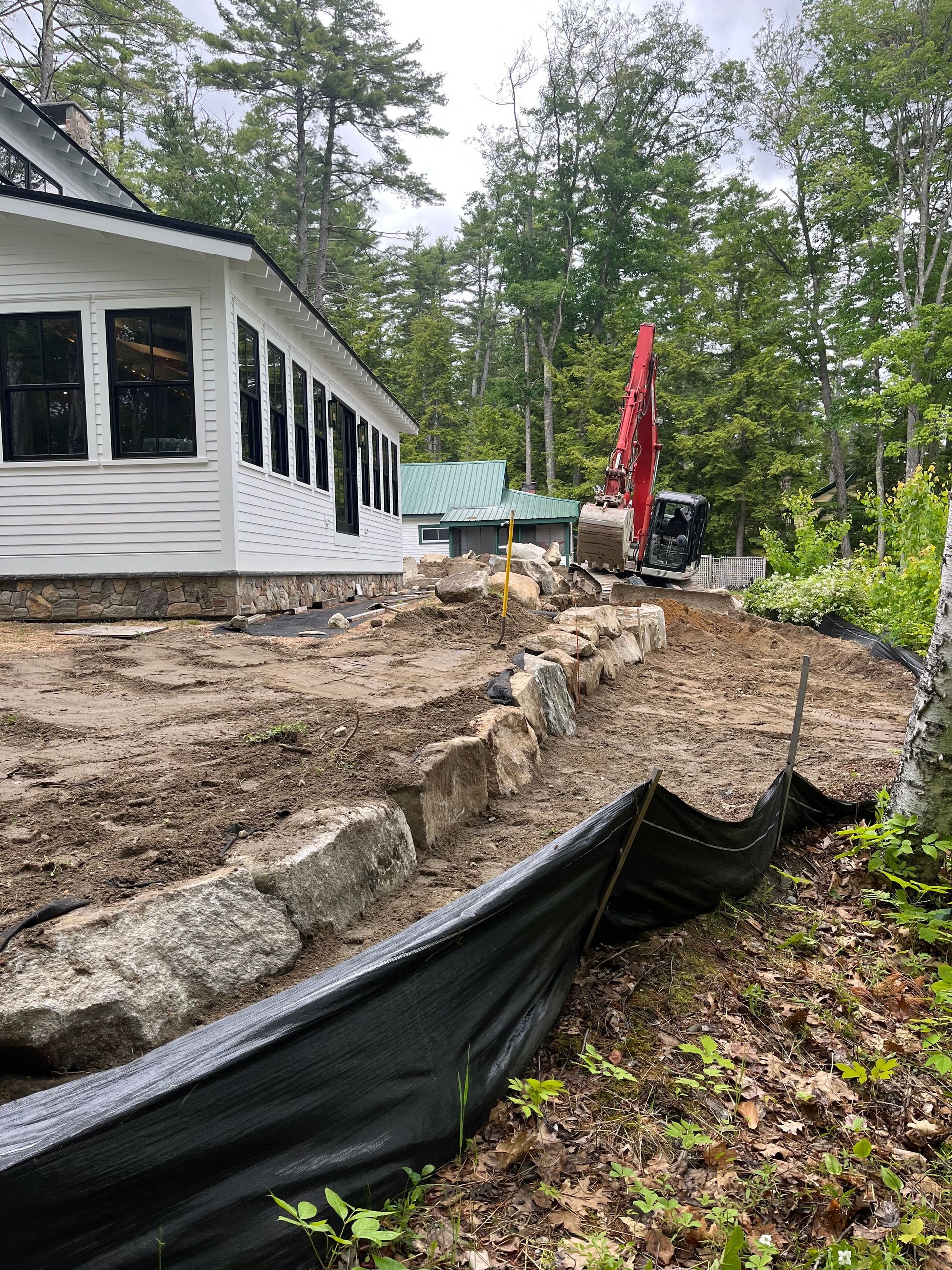 Construction equipment works near a white house with stone landscaping and black silt fencing in a wooded area.