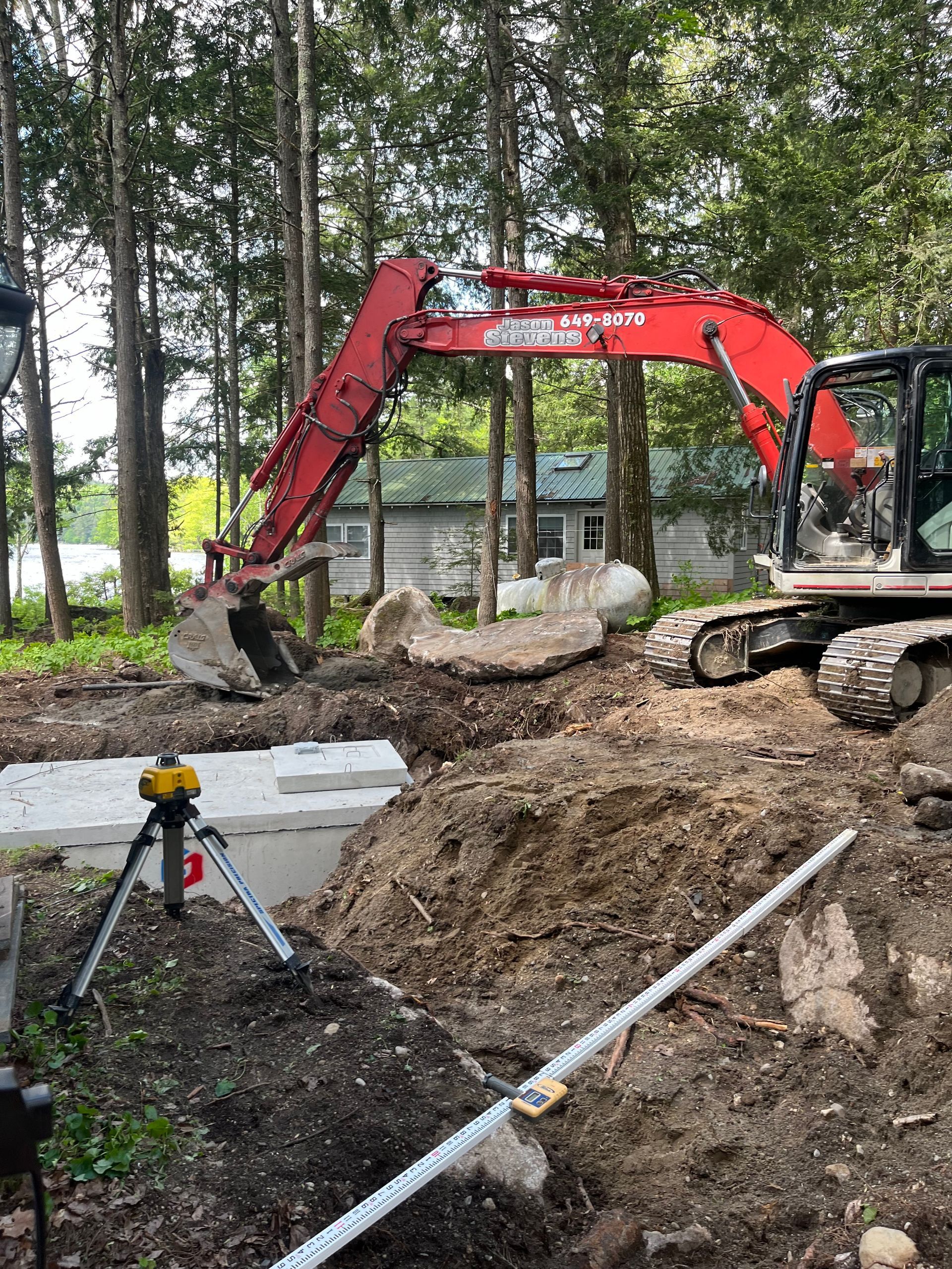 A red excavator sits near a newly installed concrete septic tank in a wooded area, with a laser level tool in the foreground.