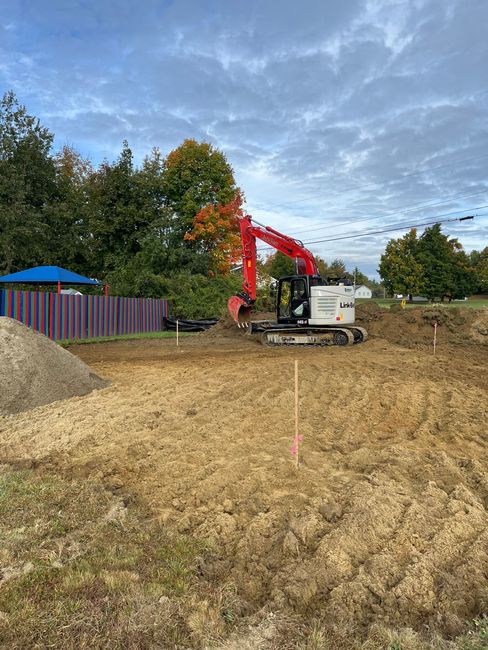 A red excavator works on a dirt construction site near a multi-colored fence and trees under a cloudy sky.