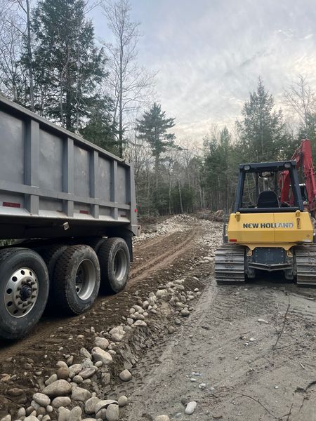 A yellow crawler loader and a large gray dump truck parked on a gravel and rocky path in a wooded area.