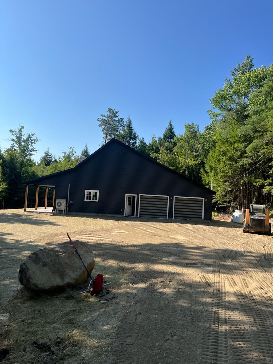 A black barn-style building sits in a clearing surrounded by woods, with a gravel yard, a large rock, and a tractor.
