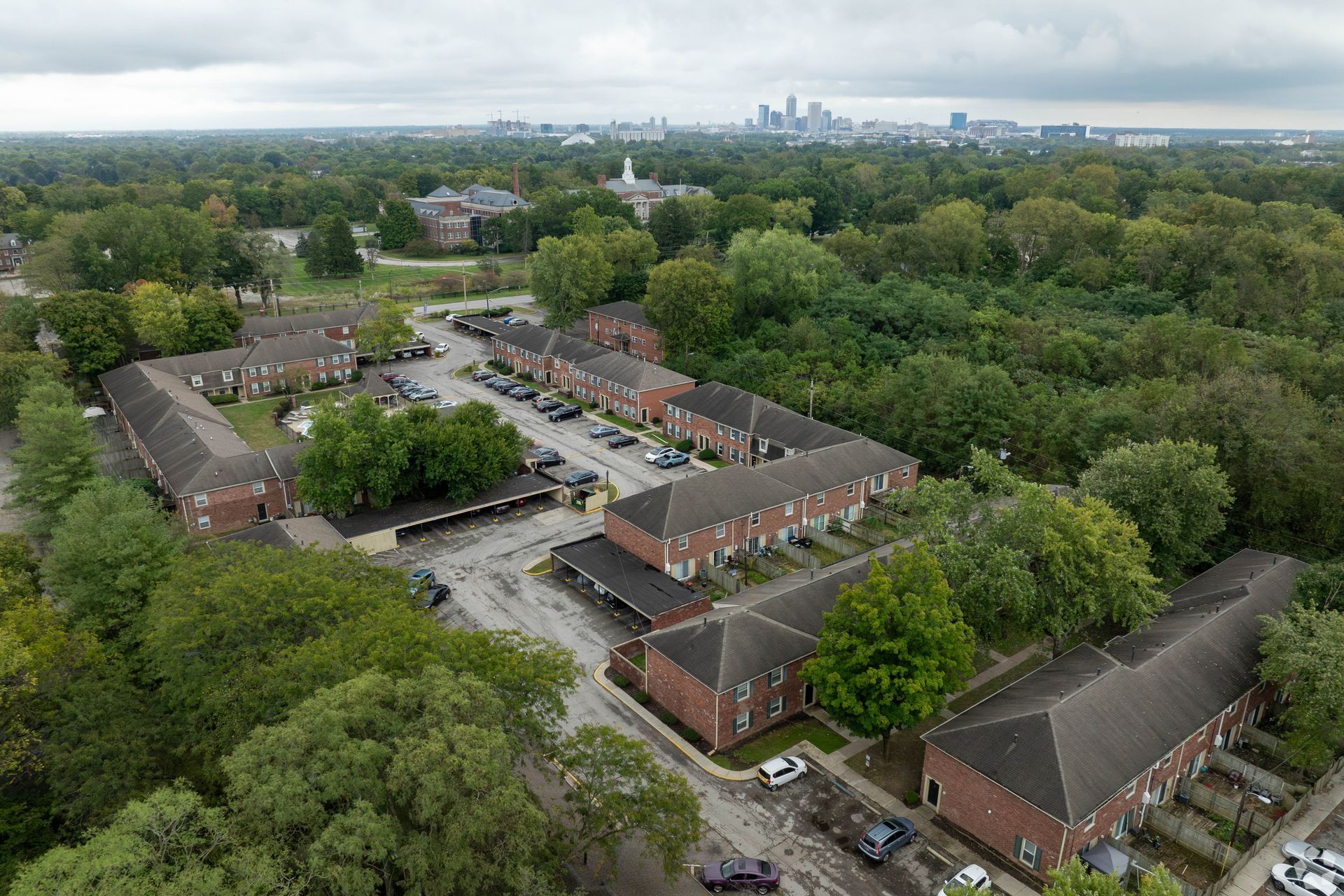 Aerial view of brick apartment buildings surrounded by trees, with a city skyline in the distance under an overcast sky.