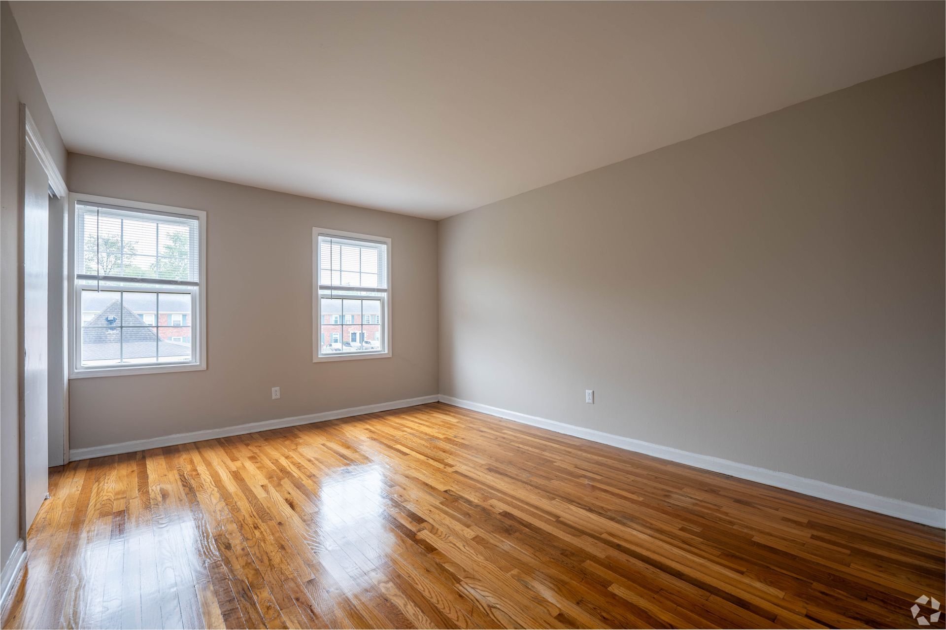 Empty room with hardwood floors, two windows, and tan walls.