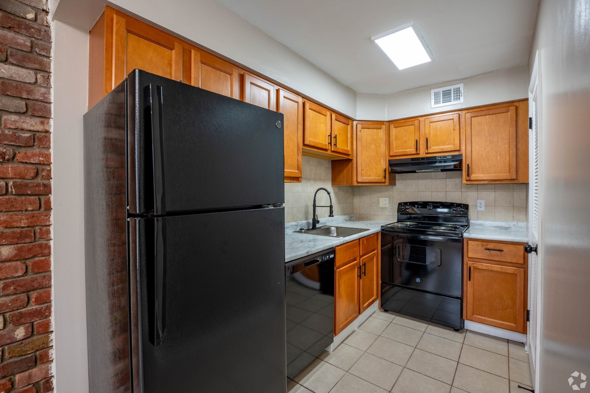 Kitchen with black appliances, wooden cabinets, brick wall, and a skylight.