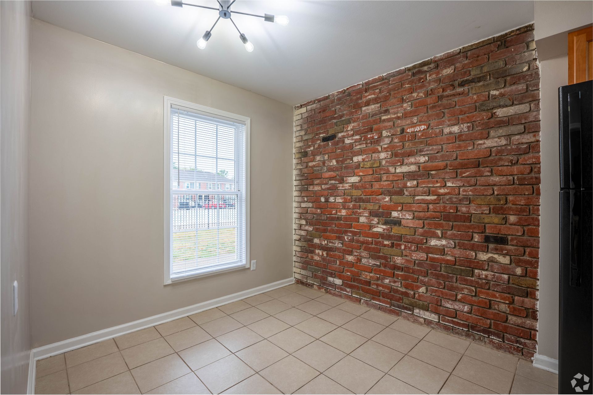 Empty room with brick accent wall, window, beige walls, tile floor, and light fixture.