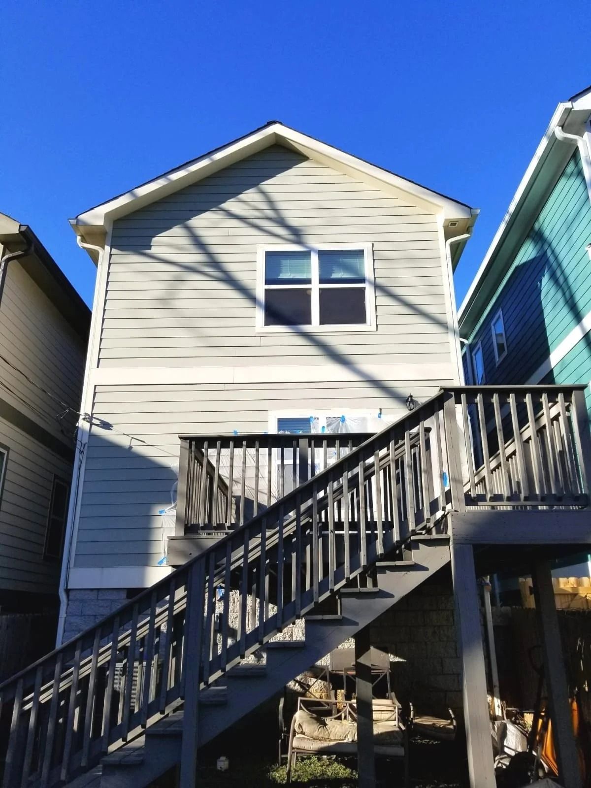 Back of a two-story house with wooden stairs leading to a deck. Gray siding, blue sky.