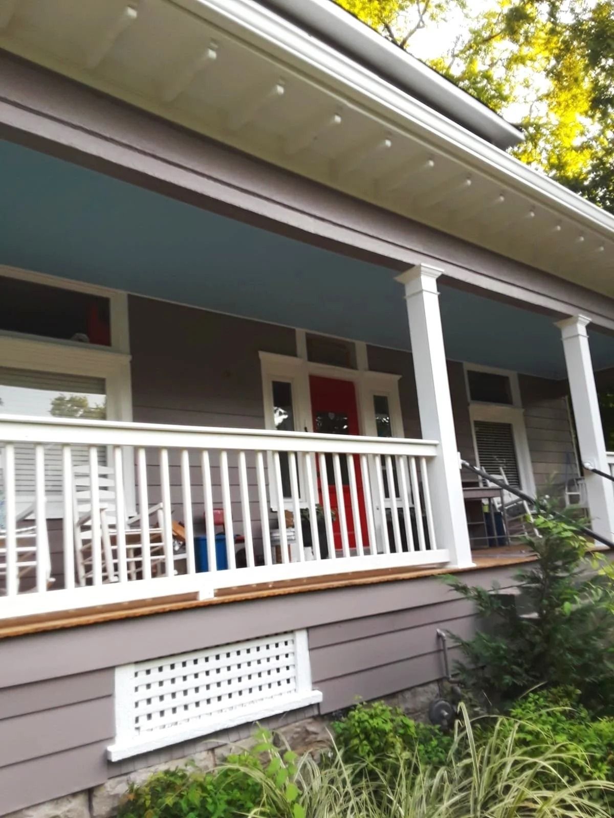 A porch with white railings, columns, and blue ceiling. Red front door. Gray house with landscaping.