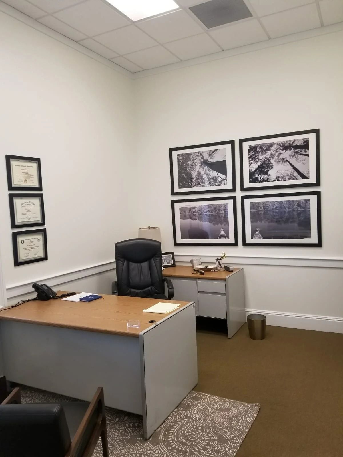 Office with desk, black chair, framed photos, and diplomas on the wall. Brown carpet and beige walls.