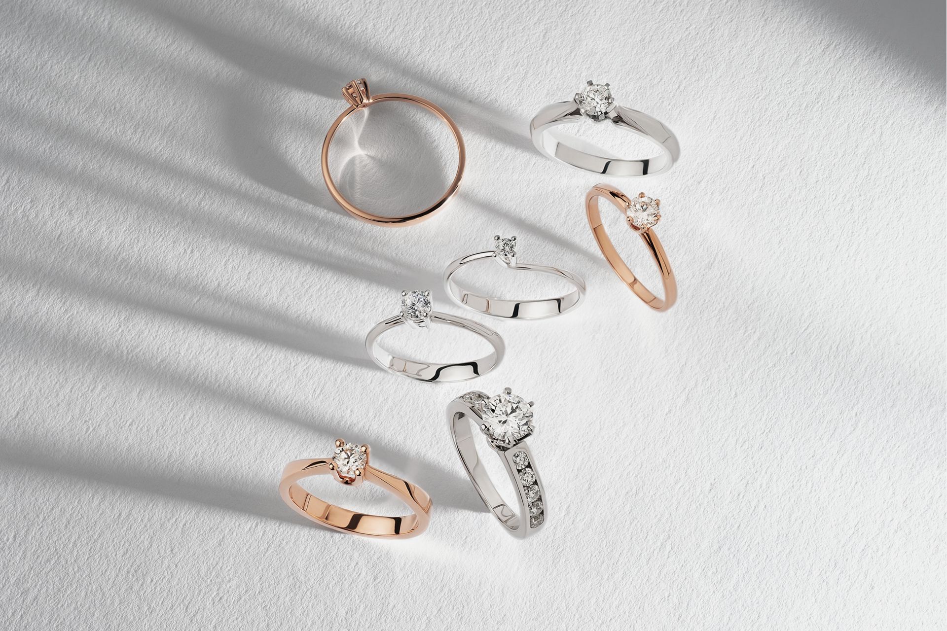 Assorted rings in rose gold, silver, and white, arranged on a light textured surface with soft shadows.