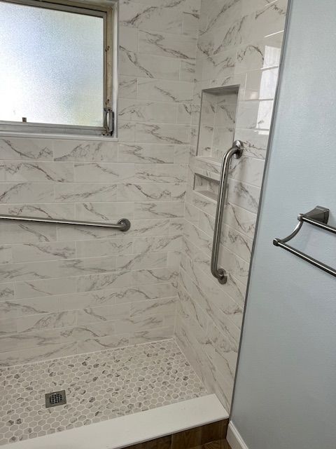 A renovated shower with white marble-look tiles, grab bars, and a pebble-stone floor.