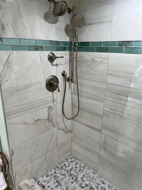 Shower stall with marble-like white and gray tiles, teal trim, pebble floor, and silver fixtures.