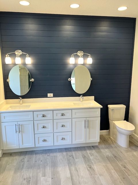 Bathroom with white vanity, blue shiplap wall, oval mirrors, and white toilet.