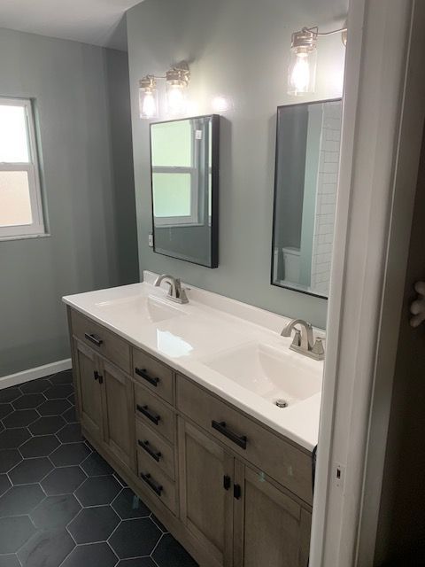 Bathroom with double sink vanity, gray walls, hexagon tile floor, and mirrors with lights.
