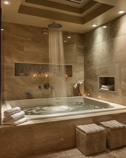 Luxurious bathroom with a bathtub under a rain shower; beige tile, towels, and two ottomans.