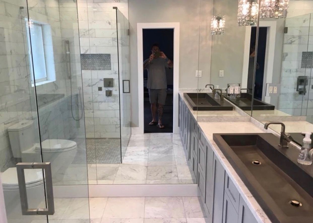 Modern bathroom with marble tile, glass shower, double sinks, and a person reflected in the mirror.