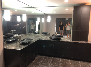 Modern bathroom with dark granite countertop, black sinks, large mirror, and two people reflected.