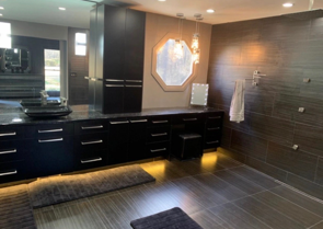 Modern bathroom with dark cabinets, tile, and octagonal mirror. Warm lighting.