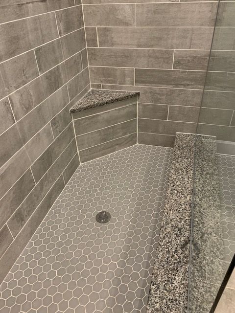 Gray tiled shower with corner seat and honeycomb floor. Glass door on the right.