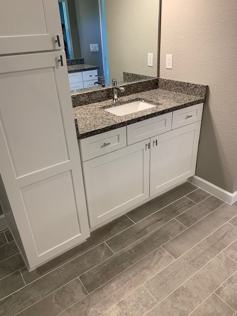 White bathroom vanity with granite countertop and mirror, next to a tall cabinet, over tile floor.