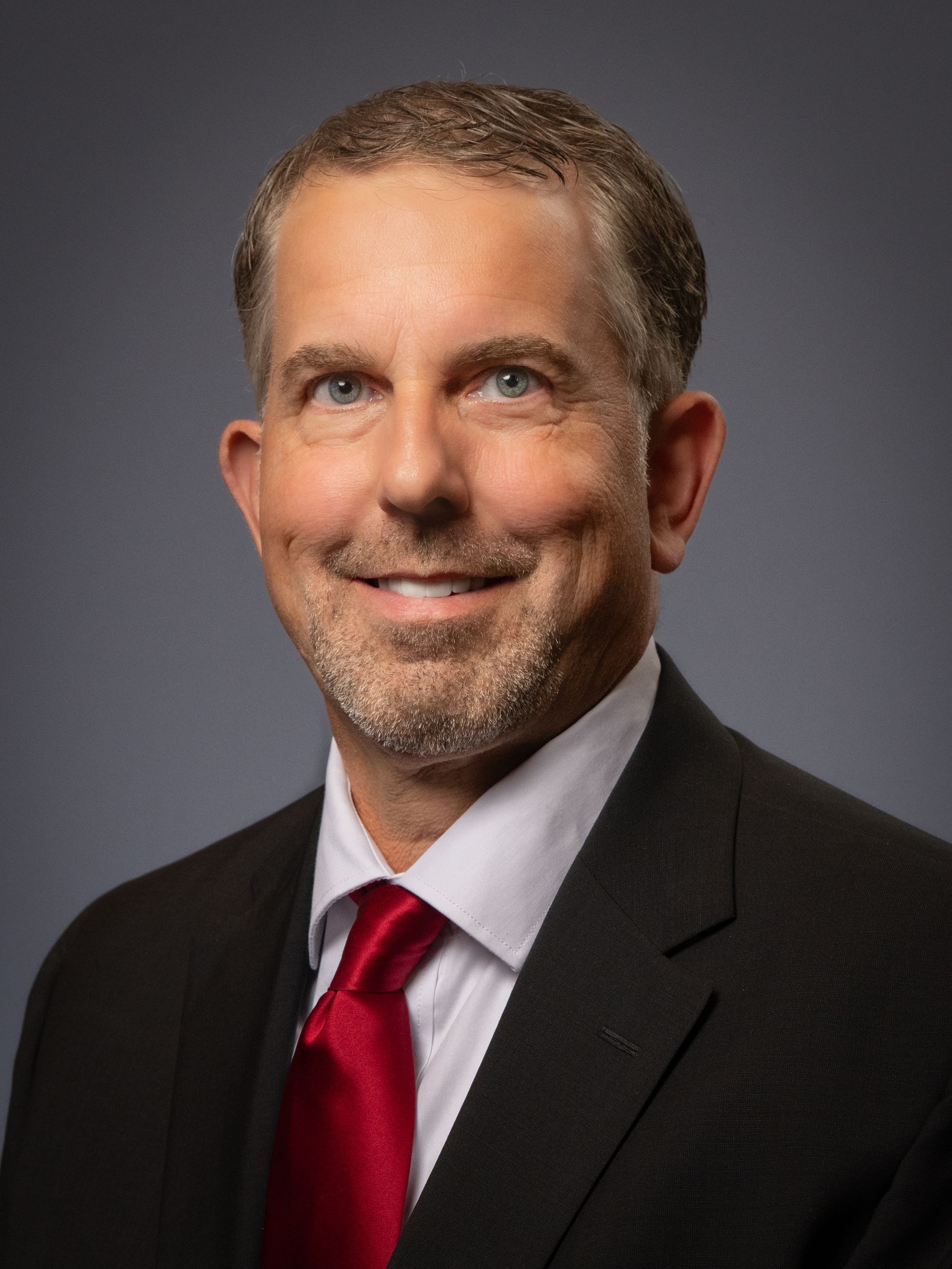 Professional headshot of a smiling man in a dark suit, white shirt, and red tie, set against a plain gray background.