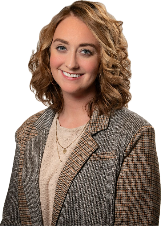 A smiling person with shoulder-length wavy blonde hair wears a patterned blazer over a beige top with a layered necklace.