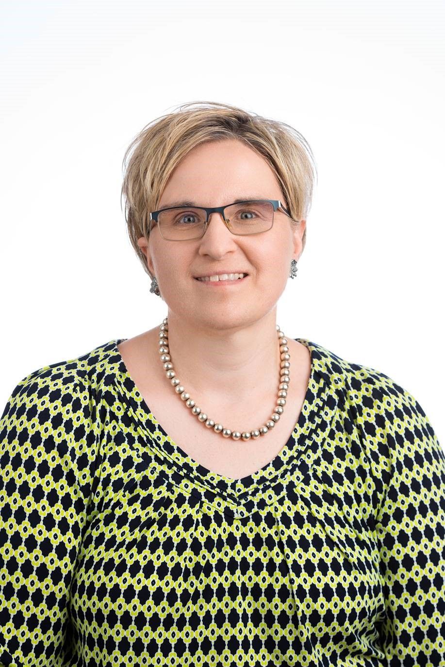 A person with short, light-brown hair wearing glasses and a lime-green and black patterned shirt with a beaded necklace.