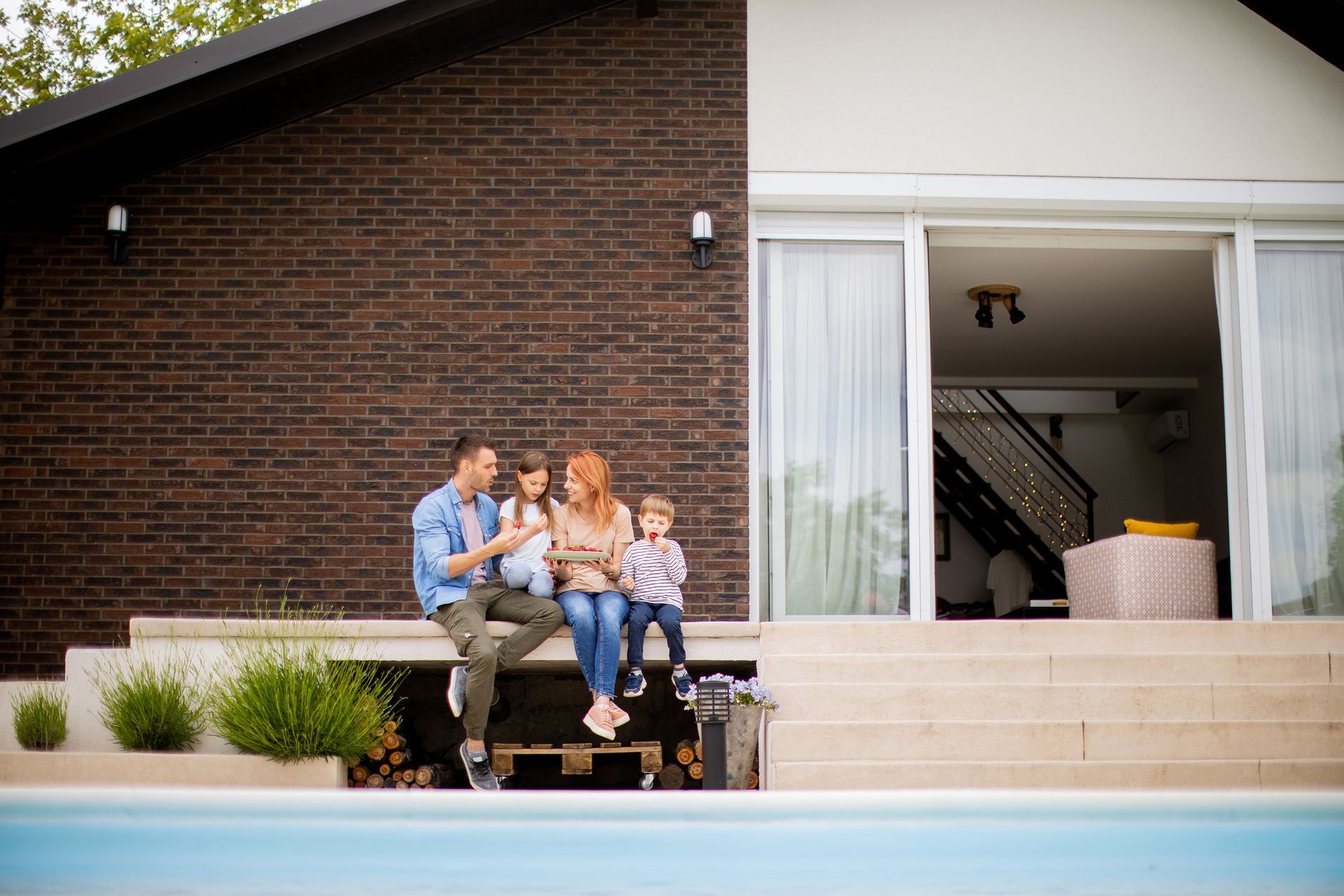 A family sits together on a patio ledge by a swimming pool in front of a modern brick home, looking at a tablet.