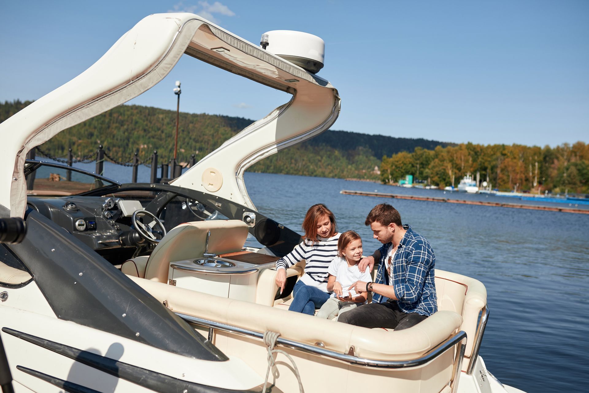 A family sits together on a boat docked in a lake, with wooded hills in the background under a clear blue sky.